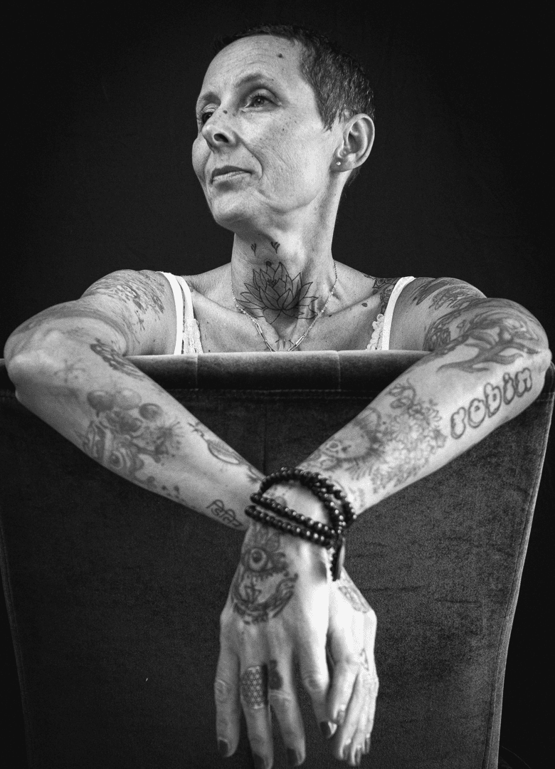 Portrait noir et blanc d'une femme tatouée accoudée au dossier d'une chaise, regard pensif