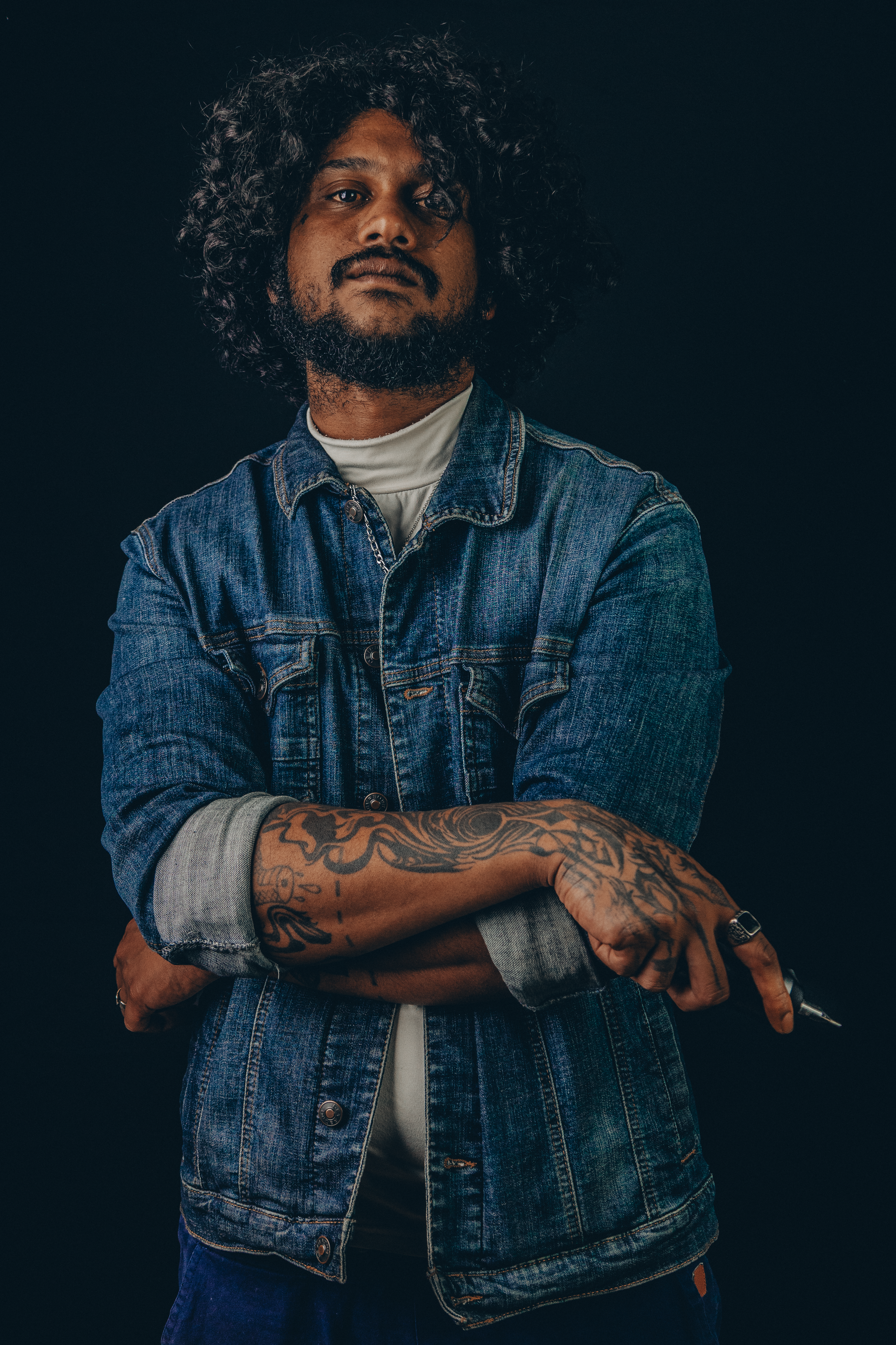 Portrait d'un homme en veste en jean, bras croisés, avant-bras tatoué, fond sombre