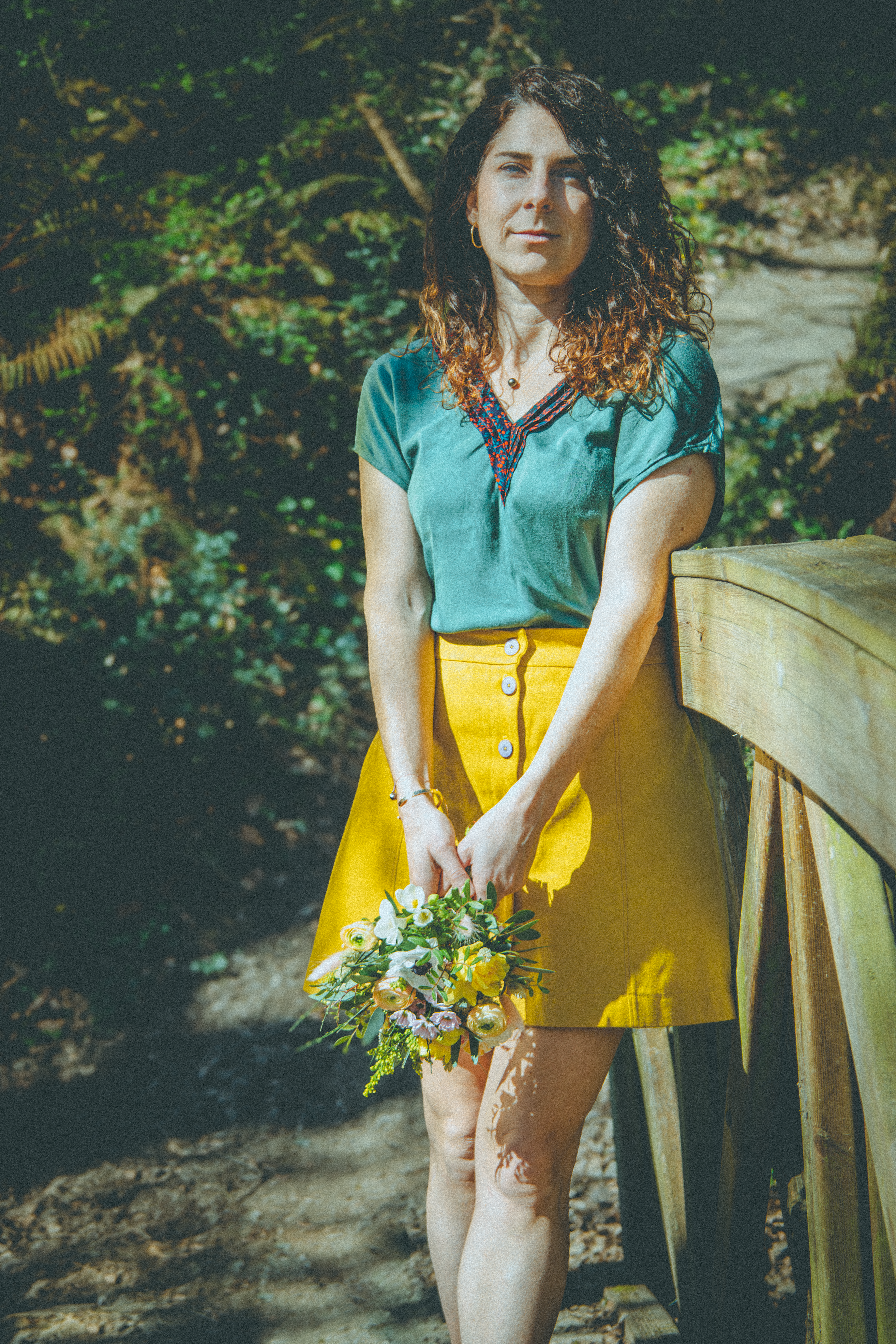 Femme en chemisier vert et jupe jaune tenant un bouquet de fleurs sur un pont en bois