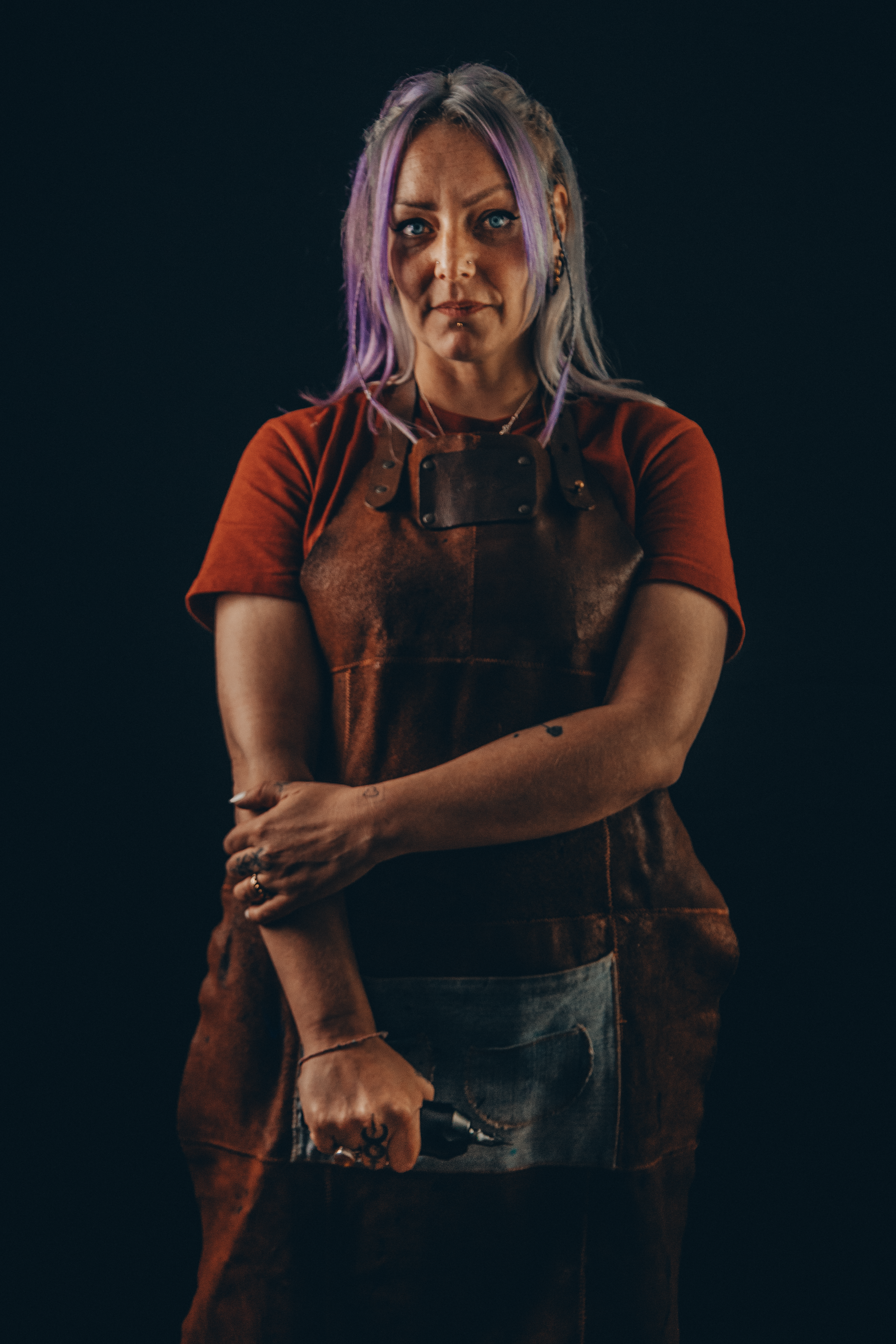 Portrait d'une tatoueuse aux cheveux violets et argentés, tablier de cuir, lumière chaude en studio